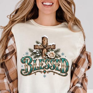 May include: A cream-coloured t-shirt featuring a graphic of a wooden cross covered in snow, accented with pine branches and a white rose. Below the cross, the word BLESSED is printed in a large, decorative teal font with brown edges.