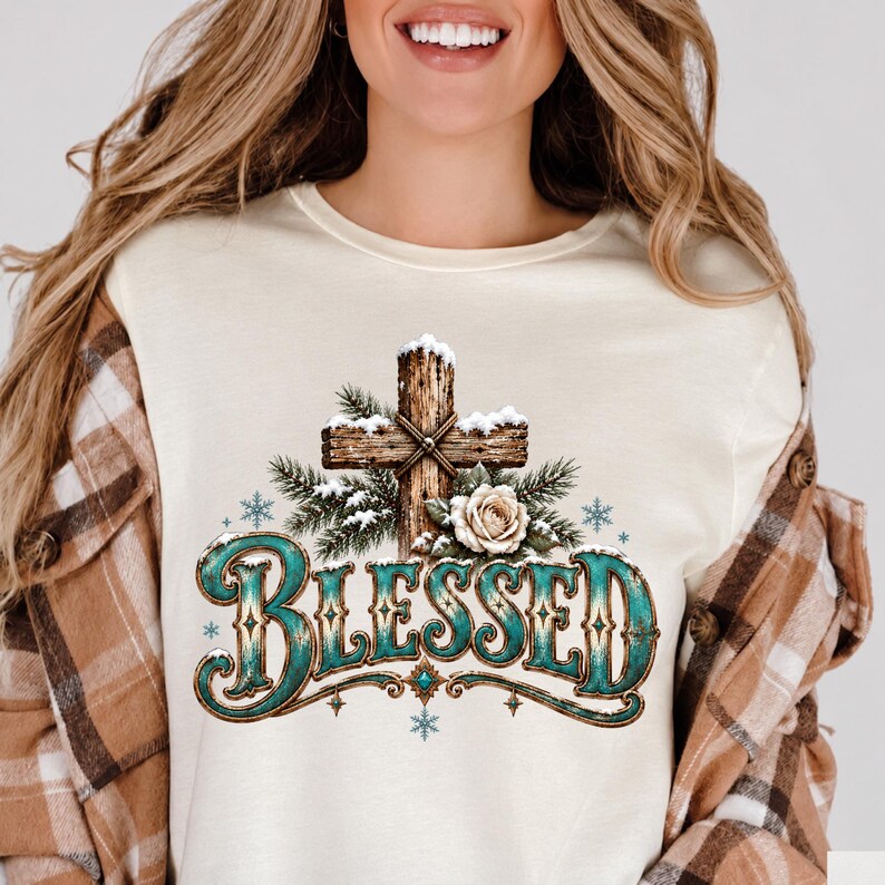 May include: A cream-coloured t-shirt featuring a graphic of a wooden cross covered in snow, accented with pine branches and a white rose. Below the cross, the word BLESSED is printed in a large, decorative teal font with brown edges.