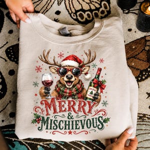 Peut inclure: Un sweat-shirt beige &agrave; col rond orn&eacute; d'un motif de renne portant un bonnet de P&egrave;re No&euml;l, des lunettes de soleil et une &eacute;charpe &agrave; carreaux. Le renne tient un verre de vin et une bouteille &eacute;tiquet&eacute;e Holiday Cheer. Le texte Merry & Mischievous est imprim&eacute;.