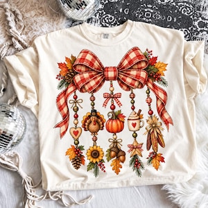 以下が含まれることがあります： 赤と白のチェック柄の大きなリボンが描かれたクリーム色のTシャツ。リボンからは、七面鳥、カボチャ、ハートが描かれたマグカップ、ヒマワリ、どんぐり、トウモロコシ、秋の葉など、秋をテーマにした様々なオーナメントがぶら下がっています。
