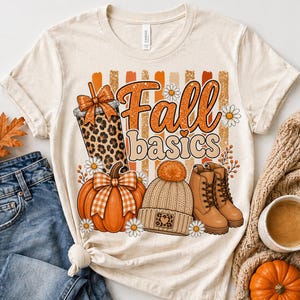 May include: Cream-colored t-shirt with the words "Fall basics" in orange and white. The design includes a pumpkin, a leopard print drink, a knit hat, boots, and daisies. A pair of blue jeans and a sweater are next to the shirt.