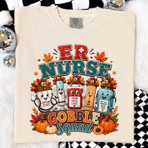 May include: A cream-colored t-shirt featuring a graphic with the text ER NURSE GOBBLE SQUAD. The design includes cartoon characters like a coffee cup, syringe, blood bag, bandage, and medicine bottle, all wearing turkey hats, surrounded by autumn leaves.