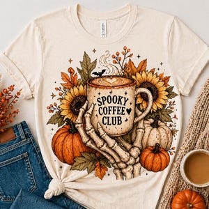 May include: Cream-colored t-shirt featuring a Halloween-themed design. The graphic includes a coffee mug with "Spooky Coffee Club" text, held by a skeleton hand, surrounded by pumpkins, sunflowers, and fall foliage.