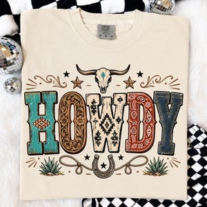 May include: A cream-colored t-shirt featuring the word HOWDY in large, patterned letters. The design includes a steer skull, stars, a horseshoe, and agave plants. Each letter has a unique texture, including turquoise, floral, geometric, paisley, and denim patterns.