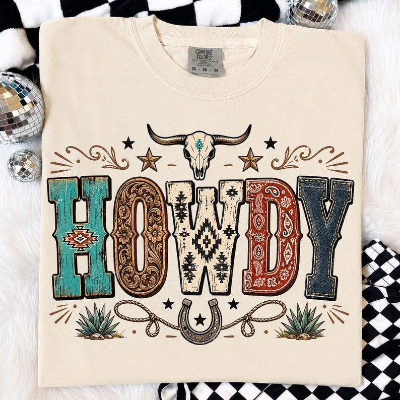 May include: A cream-colored t-shirt featuring the word HOWDY in large, patterned letters. The design includes a steer skull, stars, a horseshoe, and agave plants. Each letter has a unique texture, including turquoise, floral, geometric, paisley, and denim patterns.