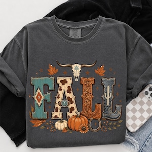 May include: A charcoal gray crewneck t-shirt featuring a graphic print that spells the word FALL. The letters are decorated with turquoise, cow print, floral leather, and denim textures. A bull skull, pumpkins, and leaves are included.