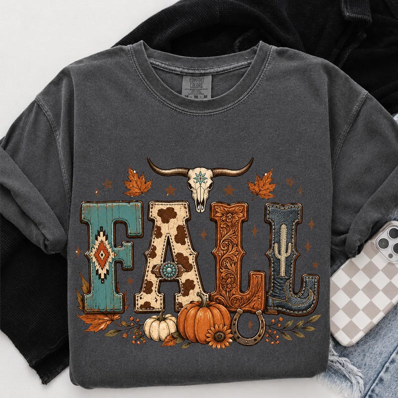 May include: A charcoal gray crewneck t-shirt featuring a graphic print that spells the word FALL. The letters are decorated with turquoise, cow print, floral leather, and denim textures. A bull skull, pumpkins, and leaves are included.