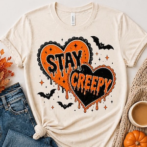May include: Cream-colored t-shirt featuring a Halloween-themed design. The graphic includes two heart shapes, one orange and one black, with the words "STAY CREEPY" in dripping text. Bats and stars complete the spooky design.