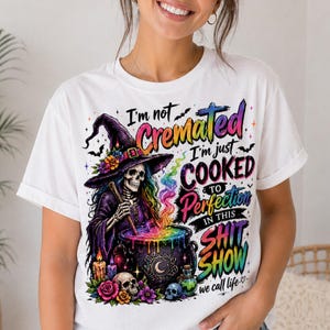 May include: White t-shirt with a colorful graphic featuring a skeleton witch stirring a cauldron. The text reads "I'm not cremated, I'm just cooked to perfection in this shit show we call life." The design includes bats, skulls, and flowers.