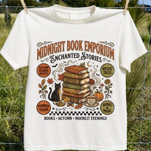 May include: A cream-colored t-shirt featuring a graphic with the text Midnight Book Emporium Enchanted Stories. The design includes a stack of books, a black cat, a candle, a cup of tea, a pumpkin, and autumn leaves with the text Books Autumn Moonlit Evenings.