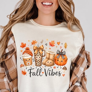 May include: A cream-coloured t-shirt featuring a graphic print of a coffee cup with an orange beanie, a leopard print tumbler with a gold bow, tan boots, and a pumpkin with a black and white bow, all with faces. The text reads Fall Vibes.