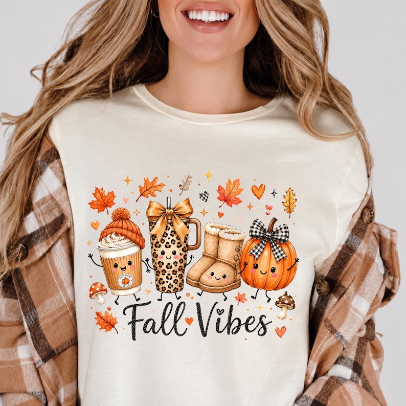 May include: A cream-coloured t-shirt featuring a graphic print of a coffee cup with an orange beanie, a leopard print tumbler with a gold bow, tan boots, and a pumpkin with a black and white bow, all with faces. The text reads Fall Vibes.