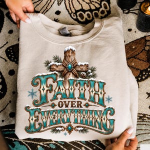 May include: A beige crewneck sweatshirt featuring a graphic print of a wooden cross covered in snow and pine branches. The text "FAITH OVER EVERYTHING" is printed in a large, decorative turquoise and brown font with snowflake accents.