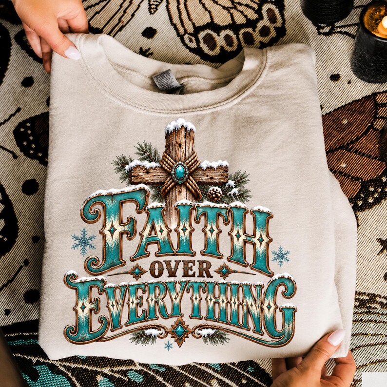 May include: A beige crewneck sweatshirt featuring a graphic print of a wooden cross covered in snow and pine branches. The text "FAITH OVER EVERYTHING" is printed in a large, decorative turquoise and brown font with snowflake accents.