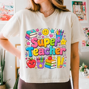 May include: A cream-colored t-shirt featuring a colorful graphic with the text Super Teacher. The design includes illustrations of stacked books, a smiling apple, a pencil holder with crayons, a ruler, a yellow star, and a smiley face.