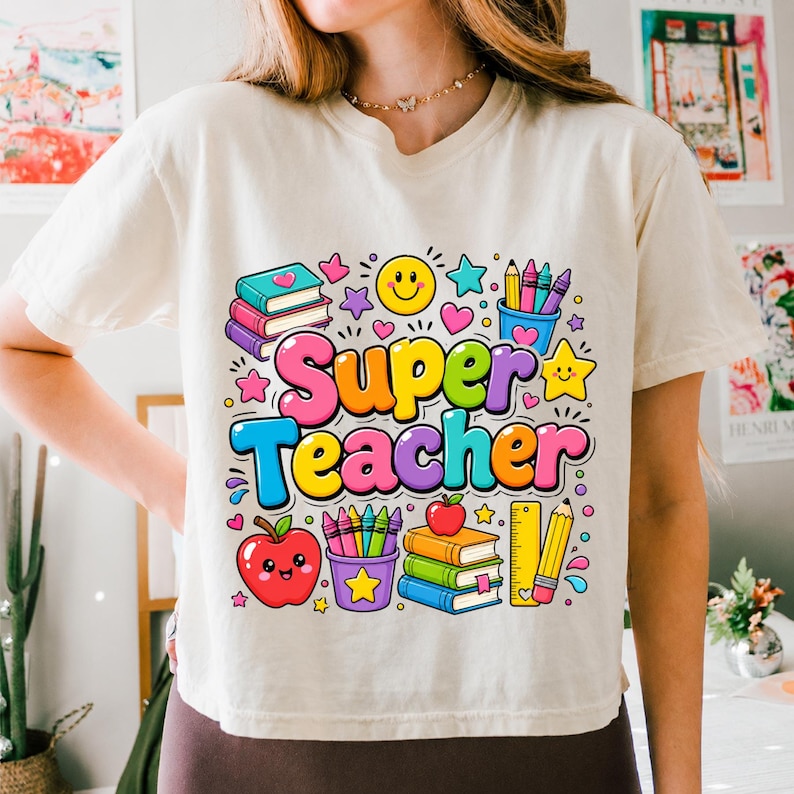 May include: A cream-colored t-shirt featuring a colorful graphic with the text Super Teacher. The design includes illustrations of stacked books, a smiling apple, a pencil holder with crayons, a ruler, a yellow star, and a smiley face.