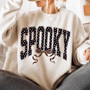 May include: A cream-coloured crewneck sweatshirt featuring the word SPOOKY in large block letters. The letters are black with white, orange, and purple polka dots. Below the text is a decorative bow with a brown and black leopard print pattern.