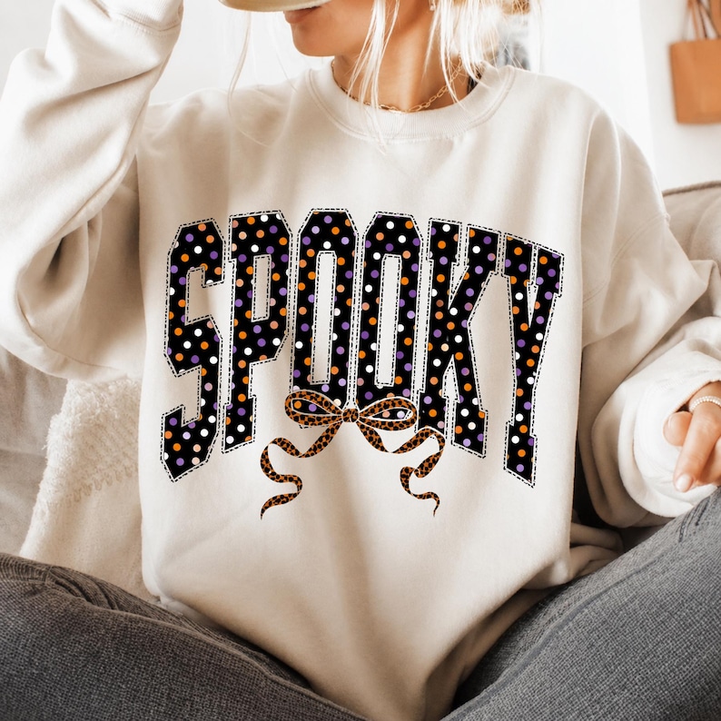 May include: A cream-coloured crewneck sweatshirt featuring the word SPOOKY in large block letters. The letters are black with white, orange, and purple polka dots. Below the text is a decorative bow with a brown and black leopard print pattern.