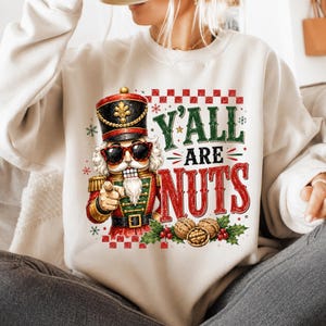 Peut inclure: Un sweat-shirt couleur cr&egrave;me avec une illustration de casse-noisette portant des lunettes de soleil et pointant du doigt. Le texte Y'ALL ARE NUTS est &eacute;crit en vert et rouge. Le motif inclut des noix, du houx et des baies avec une bordure &agrave; carreaux.