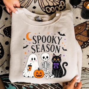 May include: A beige crewneck sweatshirt featuring a graphic print with the text "SPOOKY SEASON" in black. The design includes a white ghost holding an orange cup, a skeleton, a black cat with a purple bow, a pumpkin, bats, and stars.