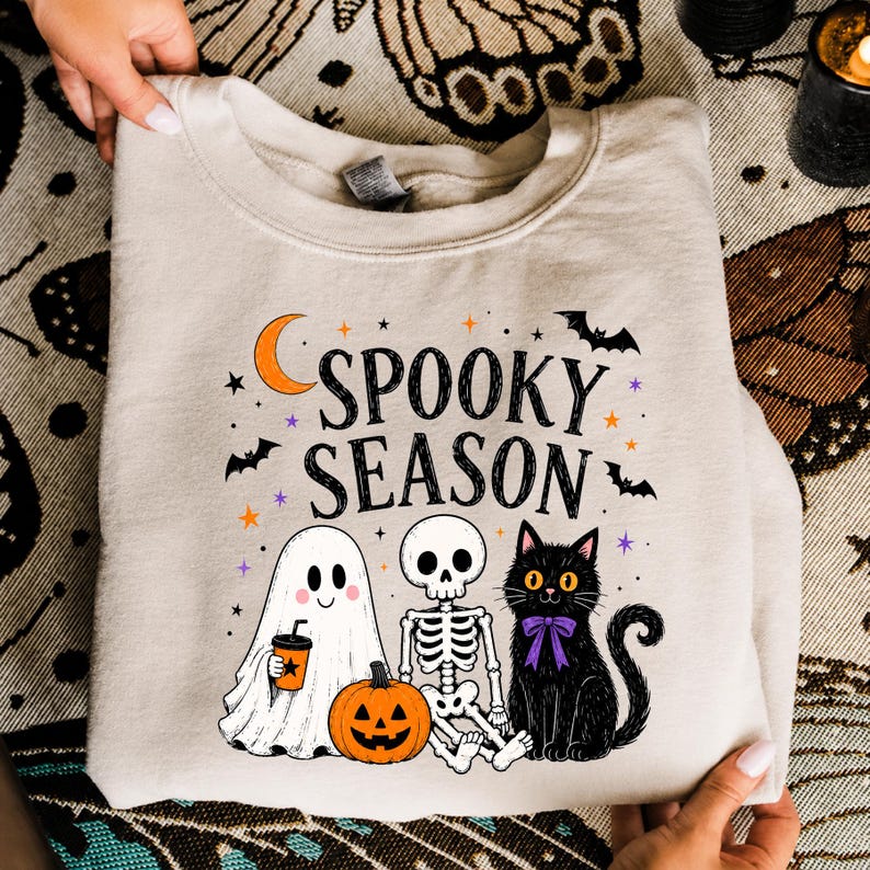 May include: A beige crewneck sweatshirt featuring a graphic print with the text "SPOOKY SEASON" in black. The design includes a white ghost holding an orange cup, a skeleton, a black cat with a purple bow, a pumpkin, bats, and stars.