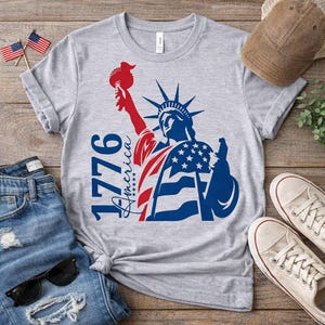 May include: A gray t-shirt featuring a patriotic design of the Statue of Liberty in red, white, and blue. The shirt includes the text "1776 America" and is paired with blue jeans, sunglasses, and sneakers.