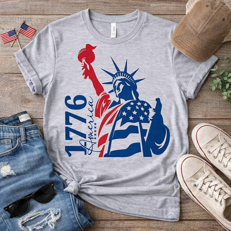 May include: A gray t-shirt featuring a patriotic design of the Statue of Liberty in red, white, and blue. The shirt includes the text "1776 America" and is paired with blue jeans, sunglasses, and sneakers.