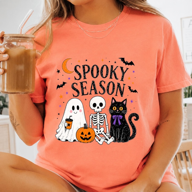May include: A coral-colored t-shirt featuring the text SPOOKY SEASON in black letters. The graphic includes a white ghost holding an orange cup, a skeleton, a jack-o'-lantern, a black cat with a purple bow, a crescent moon, and small bats.