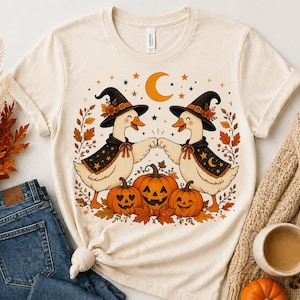 May include: Cream-colored t-shirt featuring a Halloween-themed graphic. Two cartoon geese wearing witch hats and capes bump fists, surrounded by pumpkins, leaves, stars, and a crescent moon. The shirt is paired with blue jeans and a beige sweater.