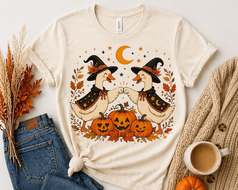May include: Cream-colored t-shirt featuring a Halloween-themed graphic. Two cartoon geese wearing witch hats and capes bump fists, surrounded by pumpkins, leaves, stars, and a crescent moon. The shirt is paired with blue jeans and a beige sweater.