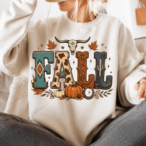 May include: A cream-colored crewneck sweatshirt featuring a graphic print of the word FALL. The letters are decorated with turquoise patterns, cow print, brown leather-style floral tooling, and denim textures, accented by a skull, pumpkins, and leaves.