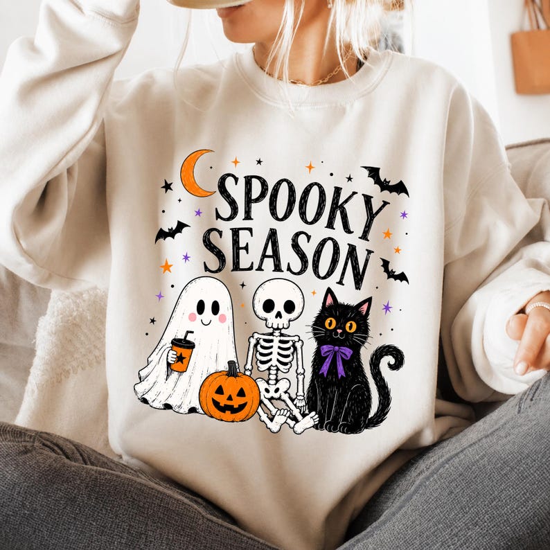 May include: A cream-colored crewneck sweatshirt featuring a graphic print with the text "SPOOKY SEASON" in black. The design includes a white ghost holding an orange cup, a skeleton, a jack-o'-lantern, a black cat with a purple bow, and bats.
