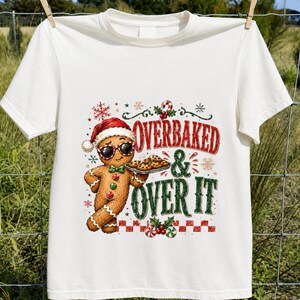 Peut inclure: Un t-shirt &agrave; manches courtes couleur cr&egrave;me avec un motif repr&eacute;sentant un bonhomme en pain d'&eacute;pice portant un bonnet de P&egrave;re No&euml;l et des lunettes de soleil, tenant un plateau de biscuits. Le texte indique OVERBAKED AND OVER IT avec des motifs festifs.