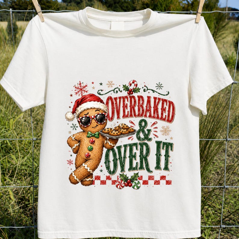 Peut inclure: Un t-shirt &agrave; manches courtes couleur cr&egrave;me avec un motif repr&eacute;sentant un bonhomme en pain d'&eacute;pice portant un bonnet de P&egrave;re No&euml;l et des lunettes de soleil, tenant un plateau de biscuits. Le texte indique OVERBAKED AND OVER IT avec des motifs festifs.
