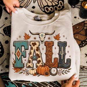 May include: A beige crewneck sweatshirt featuring a graphic print of the word FALL. The letters are decorated with turquoise patterns, cow print, brown floral carvings, and a blue denim boot. A cow skull, pumpkins, and leaves surround the text.