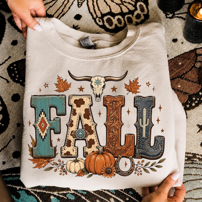 May include: A beige crewneck sweatshirt featuring a graphic print of the word FALL. The letters are decorated with turquoise patterns, cow print, brown floral carvings, and a blue denim boot. A cow skull, pumpkins, and leaves surround the text.