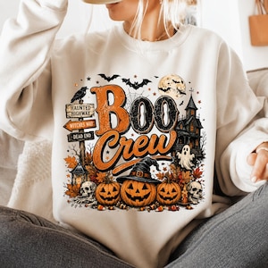 May include: A cream-colored sweatshirt featuring a graphic print with the text "Boo Crew" in orange and black. The design includes three jack-o'-lanterns, a witch hat, skulls, a haunted house, a full moon, bats, and a signpost reading Haunted Highway.