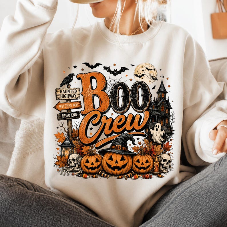 May include: A cream-colored sweatshirt featuring a graphic print with the text "Boo Crew" in orange and black. The design includes three jack-o'-lanterns, a witch hat, skulls, a haunted house, a full moon, bats, and a signpost reading Haunted Highway.