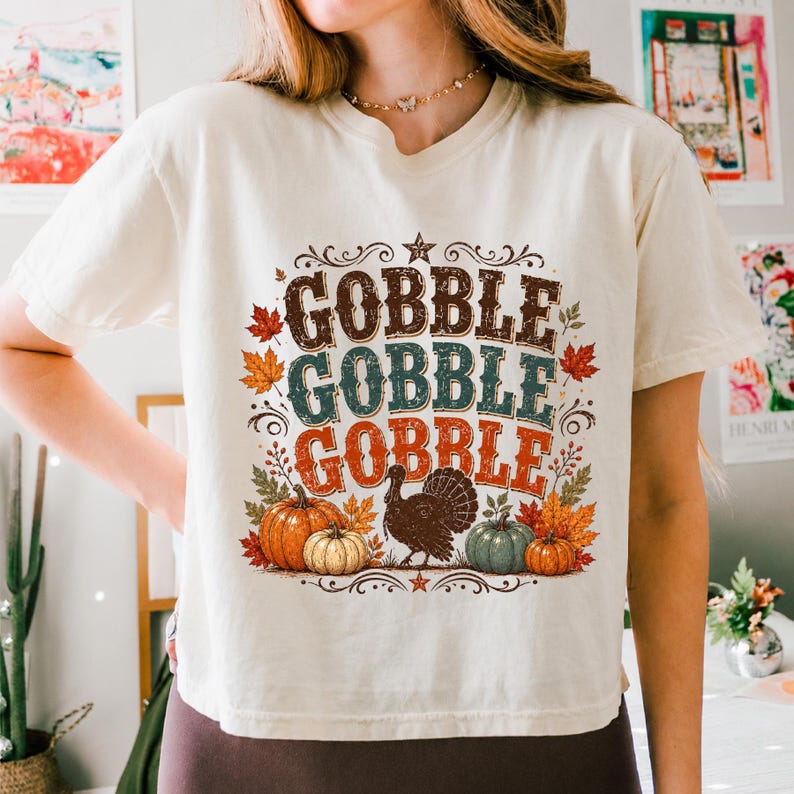 May include: A cream-colored short-sleeve t-shirt featuring a graphic print with the text GOBBLE GOBBLE GOBBLE in brown, teal, and orange. The design includes a central turkey illustration surrounded by orange, green, and red pumpkins and autumn leaves.