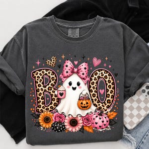 May include: A charcoal gray t-shirt featuring a graphic of a white ghost wearing a pink leopard print bow. The ghost holds a pink mug and a pumpkin bucket. The text BOO is written in large leopard print letters. Sunflowers and pink roses surround the base.
