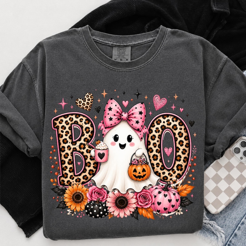 May include: A charcoal gray t-shirt featuring a graphic of a white ghost wearing a pink leopard print bow. The ghost holds a pink mug and a pumpkin bucket. The text BOO is written in large leopard print letters. Sunflowers and pink roses surround the base.