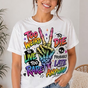 May include: White t-shirt with a colorful graphic featuring a skeleton hand making a peace sign. The text reads "Too Weird To Live Too Rare To Die Probably Late Anyway" in various colors and fonts.