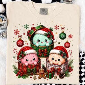 Può includere: Una t-shirt color crema con una grafica di tre creature rotonde e carine dentro scatole regalo. Le creature sono verde chiaro, rosa e beige, tutte con indosso cappelli da Babbo Natale. Il design include palline di Natale, fiocchi di neve e un omino di pan di zenzero.