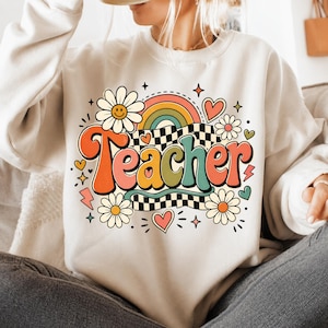 May include: A cream-colored crewneck sweatshirt featuring a colorful graphic print on the front. The design includes the word Teacher in large, bold, multi-colored letters, surrounded by daisies, a rainbow, hearts, lightning bolts, and stars.