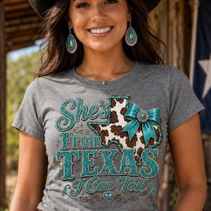 Puede incluir: Camiseta gris con el texto "She's From Texas I Can Tell" en purpurina turquesa. El dise&ntilde;o incluye un gr&aacute;fico en forma de Texas con un estampado de piel de vaca y un lazo turquesa. La modelo lleva pendientes y un collar turquesa.