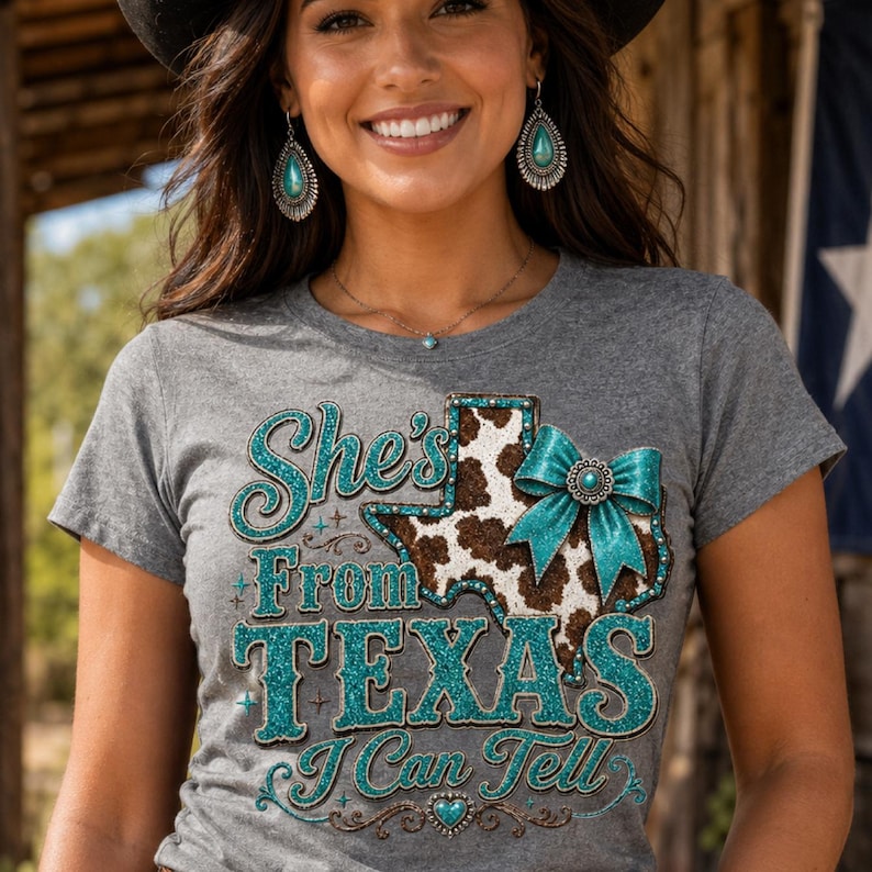 Puede incluir: Camiseta gris con el texto "She's From Texas I Can Tell" en purpurina turquesa. El dise&ntilde;o incluye un gr&aacute;fico en forma de Texas con un estampado de piel de vaca y un lazo turquesa. La modelo lleva pendientes y un collar turquesa.