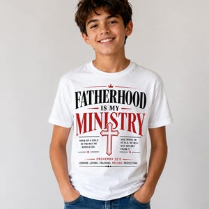 Peut inclure: T-shirt blanc avec l'inscription "FATHERHOOD IS MY MINISTRY" en noir et rouge. Le t-shirt pr&eacute;sente une croix et le verset biblique Proverbes 22:6. Le t-shirt contient &eacute;galement les mots "LEADING. LOVING. TEACHING. PRAYING. PROTECTING."