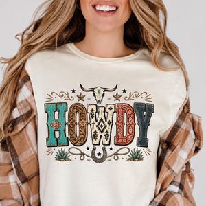 May include: A cream-colored t-shirt featuring the word HOWDY in large, patterned letters. The design includes a steer skull, stars, a horseshoe, and agave plants. Each letter has a unique texture, including turquoise, floral, geometric, and paisley patterns.