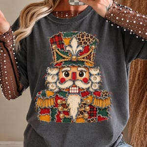 Puede incluir: Una camiseta gris carbón de manga larga con un gráfico de un cascanueces hecho de parches con patrones de cuadros, estampado de leopardo y colores sólidos. Las mangas son de malla marrón transparente adornadas con pequeñas perlas blancas.
