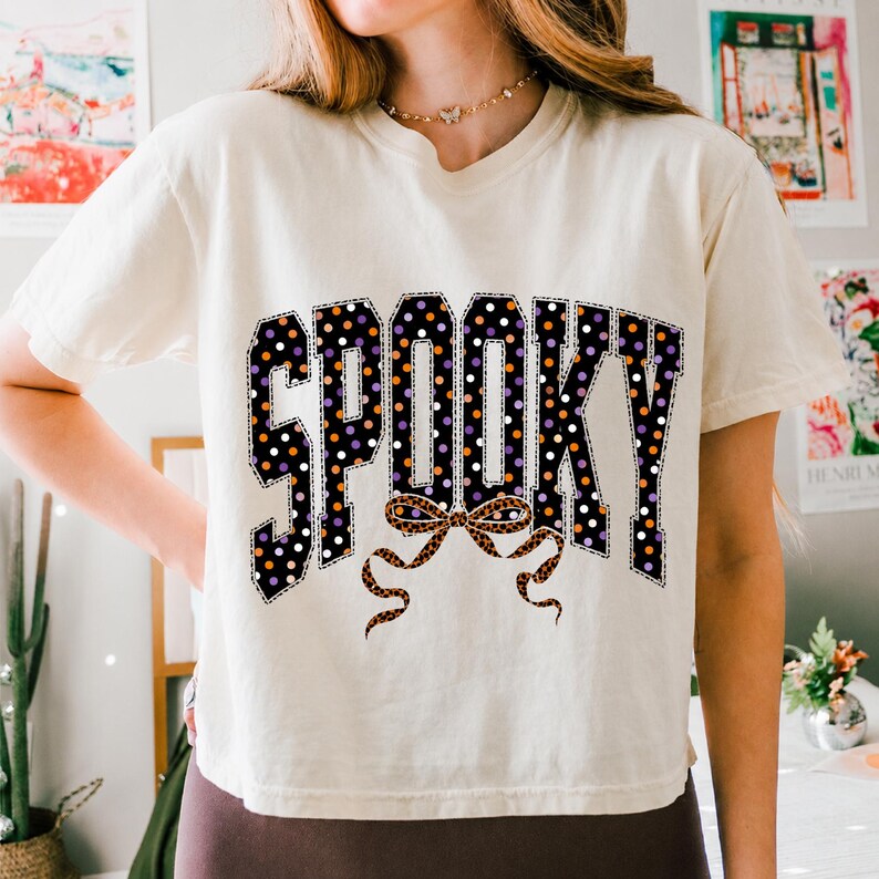 May include: A cream-coloured short-sleeve t-shirt featuring the word SPOOKY in large block letters. The letters are black with white, orange, and purple polka dots. A leopard print bow graphic is positioned directly below the word SPOOKY.
